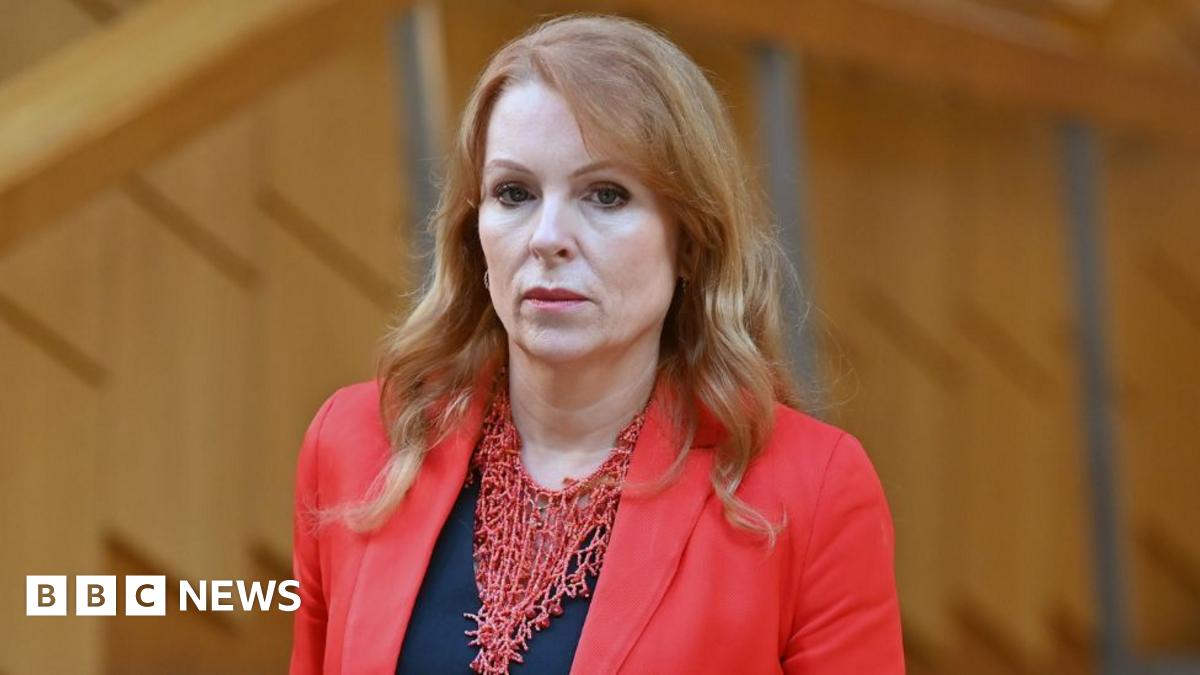 Ash Regan, who has long ginger hair, looks towards the camera, with wood panelling behind her. She is wearing a red jacket over a black top.