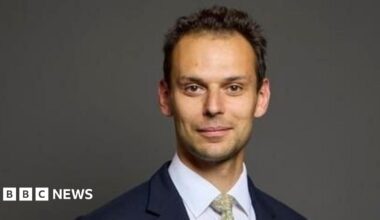 A man with short, dark, curly hair looks at the camera, wearing a navy jacket, white shirt and a pale green patterned tie.