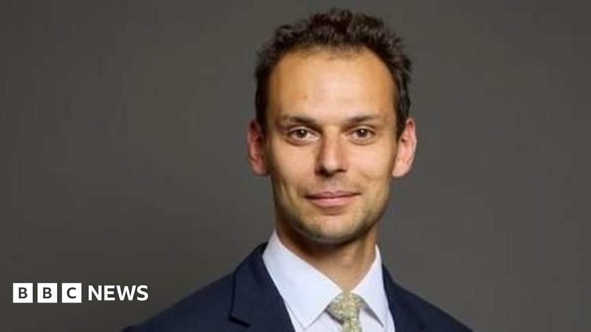 A man with short, dark, curly hair looks at the camera, wearing a navy jacket, white shirt and a pale green patterned tie.