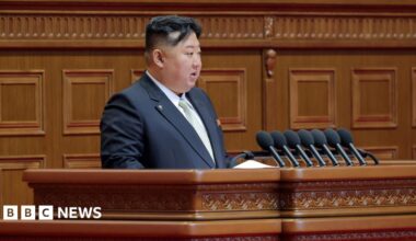Kim Jong Un speaking at a podium, wearing a black suit.