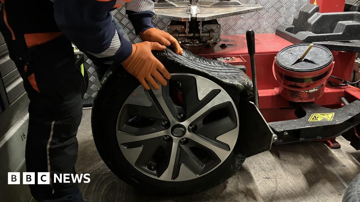 Part of the road with large, wavy cracks in the tarmac and raised sections. Orange and white cones stretch along the middle of the road as cars pass.