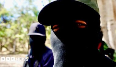 A cartel member wearing a black balaclava looks into the camera, another - also wearing a balaclava, stands in the background.