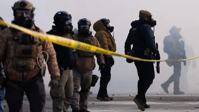 Federal agents stand behind yellow tape amid a haze caused by some form of gas. They are armed and have their faces covered