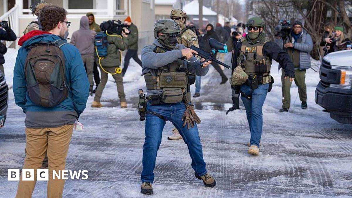 An officer with US Immigration and Customs Enforcement or ICE (right) and another federal officer holding a crowd control device (centre) stand at a Minneapolis intersection where protesters had gathered after the death of Renee Nicole Good. Bystanders and journalists also stand in the intersection.