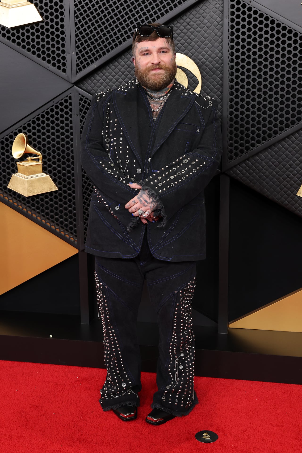 A man poses on the red carpet wearing a black studded and embellished suit with blue contrast stitching and frayed hems.