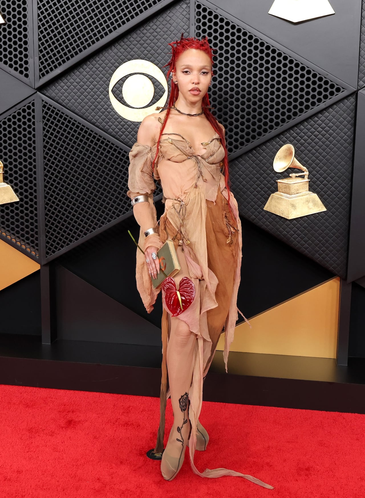 A woman poses on the red carpet, wearing a light brown gown and holding a book and a red peace lily.