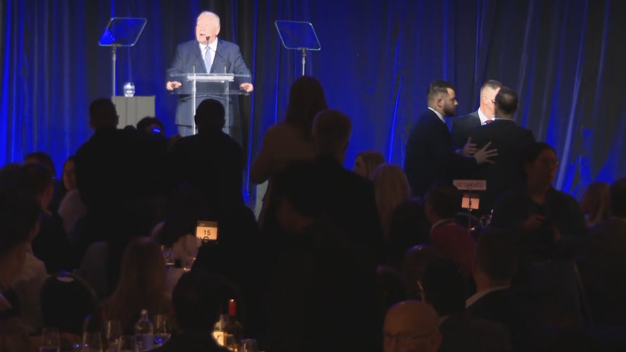 People being held and escorted out of event by security with a man at a podium in the background.