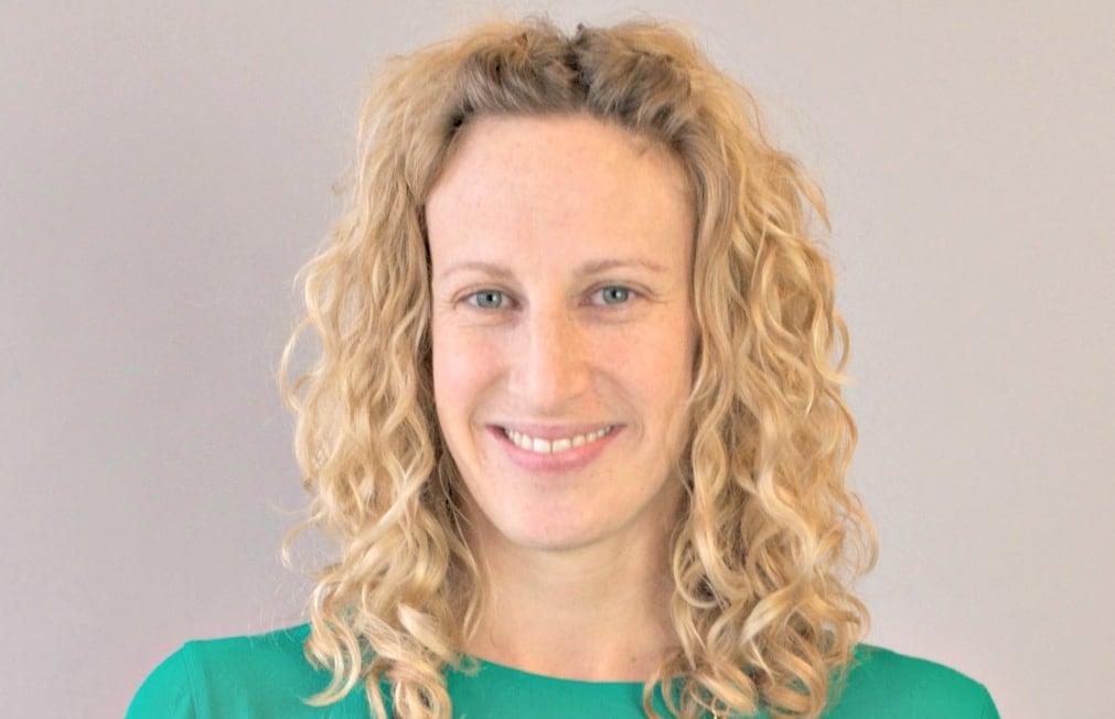 A woman with blonde curly hair, a green top and a necklace smiles.