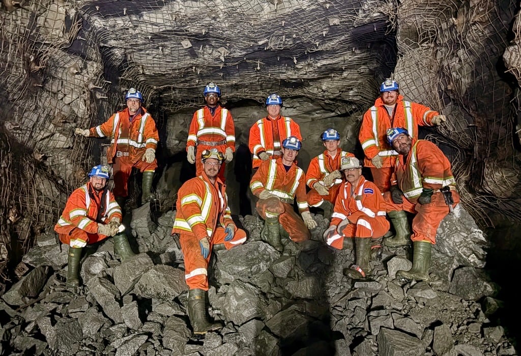 A group of student miners wearing orange coveralls stand on a large rock burst underground.