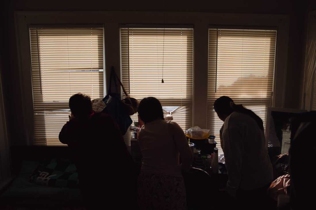 M and her oldest children peek through a window as a police car sits outside their home in Minneapolis. Jan. 26, 2026. Zaydee Sanchez for NPR