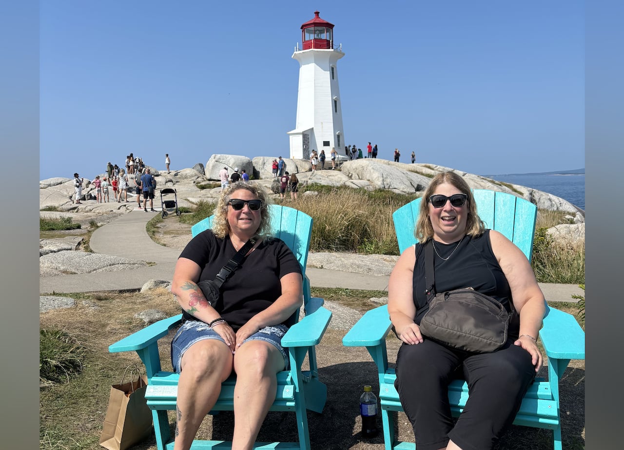 Two women with light hair sit in blue chairs in front of a red and white lighthouse