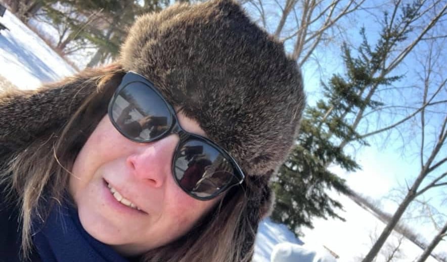 Woman with blonde hair wears a fur hat, sunglasses, blue scarf and dark winter jacket on a snowy winter day