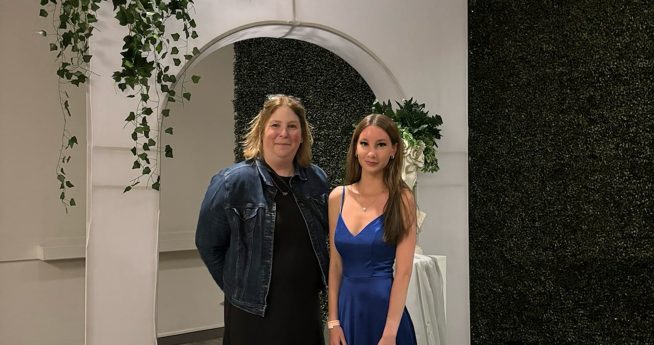 Woman with blonde hair wears a black dress and jean jacket while standing next to a young woman wearing a long blue dress in in front of a white archway