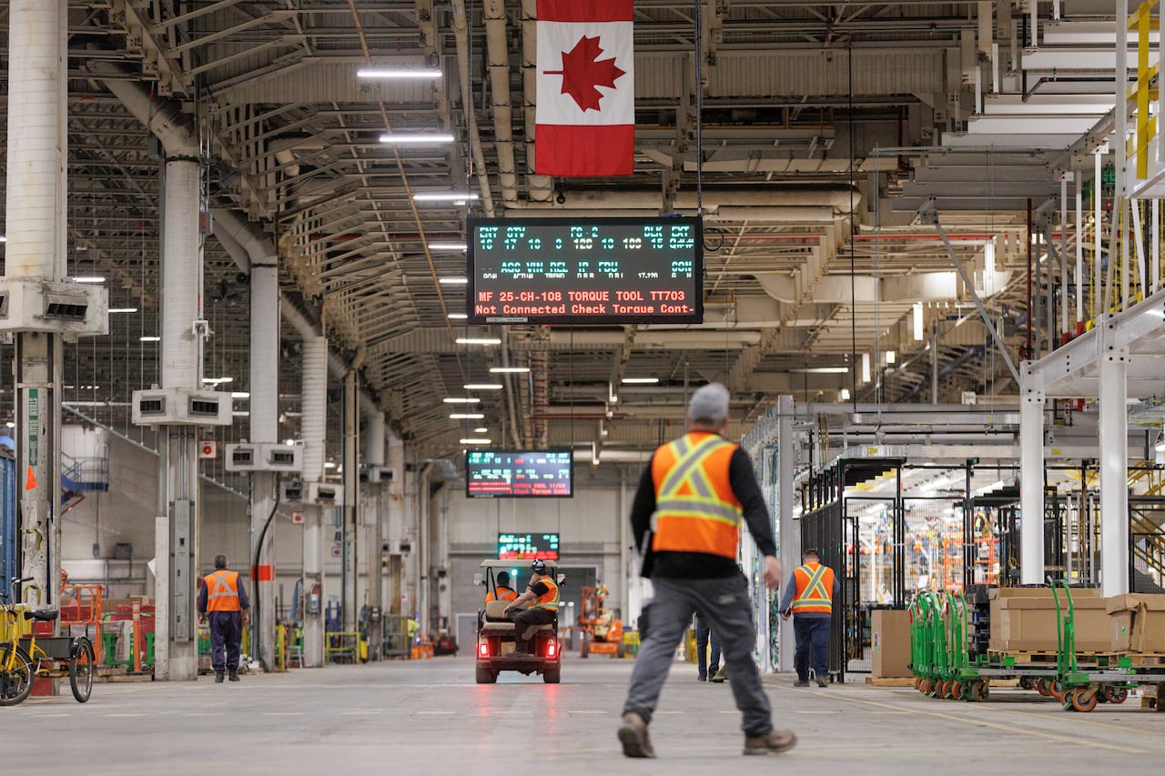 workers in orange reflective vests walk on the floor of a production plant where cars are being assembled