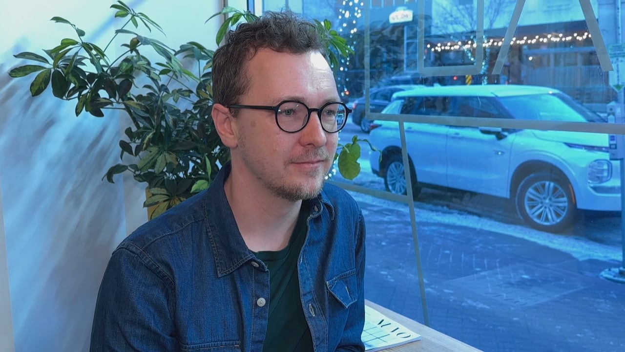 A man with brown hair and circular glasses slightly smiles while sitting down in front of a window facing a city street.