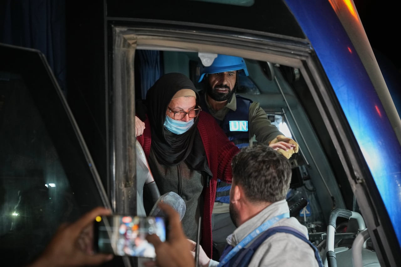 An older bespectacled woman wearing a headscarf and a medical mask over her mouth is shown being helped off a vehicle.