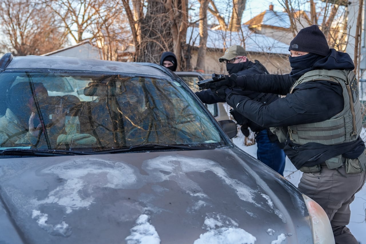 Two people are shown inside a vehicle, with at least two people in uniforms outside the vehicle pointing guns in their direction.