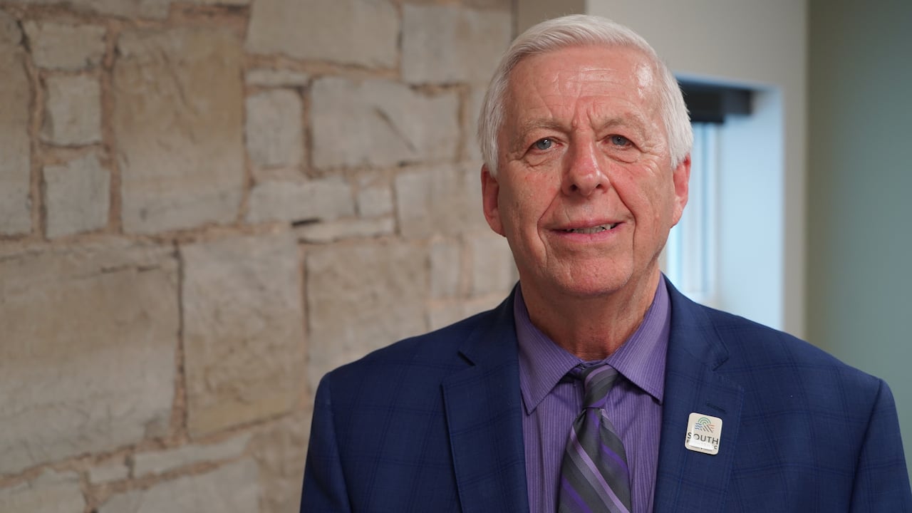 A ma with grey hair, wearing a blue suit jacket, tie and purple shirt, stands in front of a stone wall.
