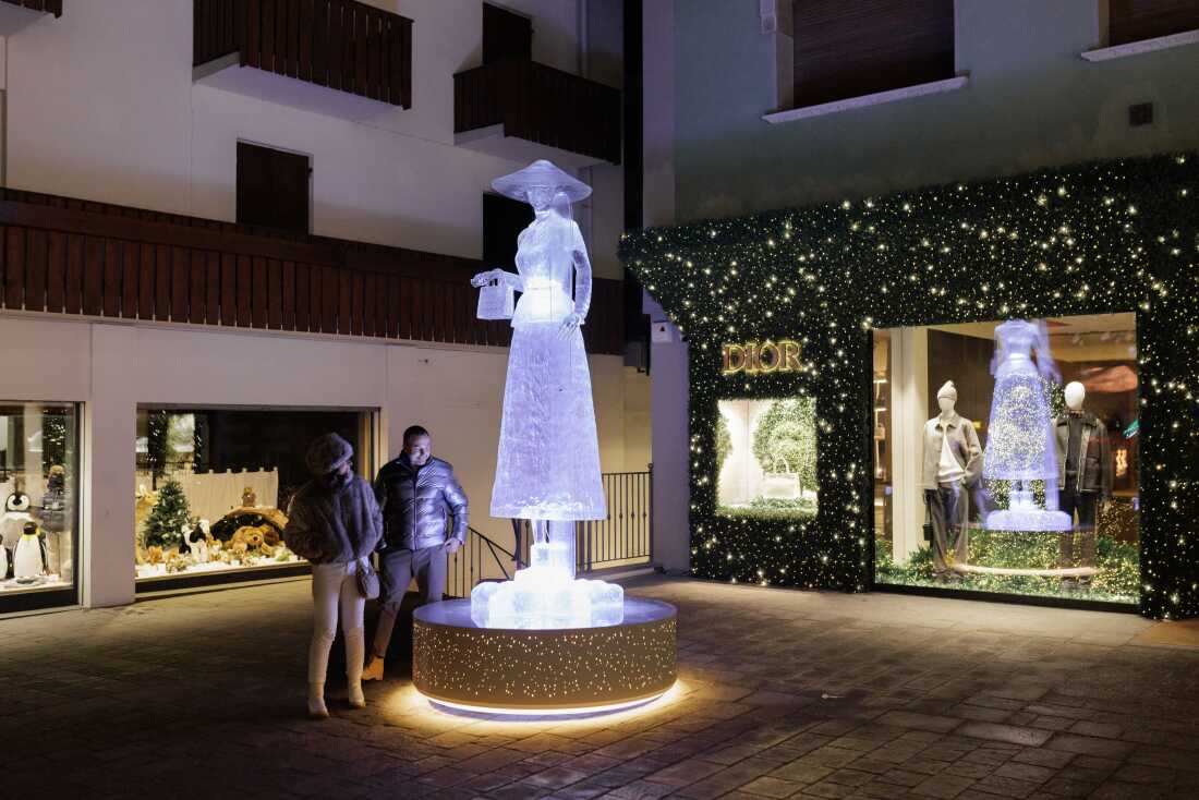 A glowing, life-sized ice sculpture of a woman in an elegant dress and hat stands illuminated at night in a pedestrian area, with the high-end Dior boutique visible in the background. As the town prepares for the Milano Cortina 2026 Winter Olympics, the presence of major global luxury brands underscores the resort's status as an elite international destination
