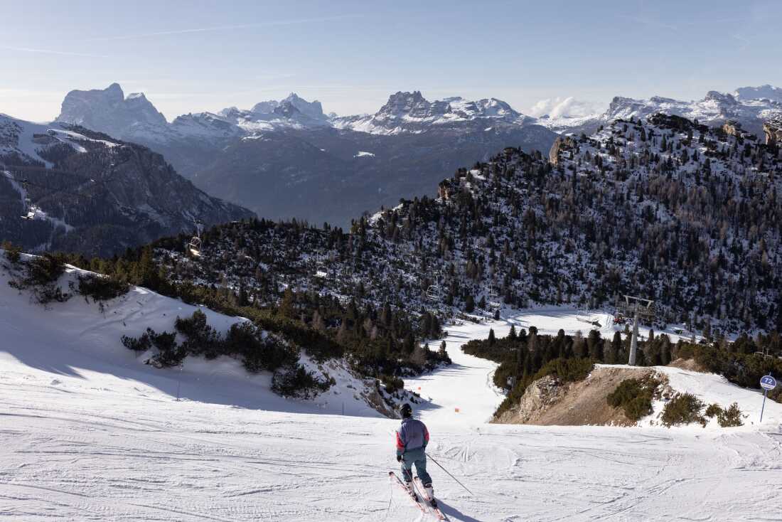A skier stands at the top of a run served by the Tre Croci - Son Forca chairlift, looking out over the Ampezzo valley before beginning a descent. The site, dominated by the Cristallo massif, highlights the intersection of high-altitude sport and the luxury branding that characterizes Cortina's preparation for the Milano Cortina 2026 Winter Olympics