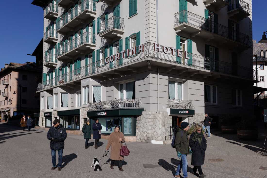 pedestrians walk past the historic Hotel Cortina and luxury retail shops in the town's central pedestrian area. As the 2026 Winter Olympics approach, the contrast between Cortina’s traditional Alpine charm and the increasing commercialization has become a focal point for local critics. While tourists enjoy the scenic town center, environmental and civic groups warn that the massive influx of Olympic-related infrastructure is shifting the town’s identity toward a more urbanized, high-density model