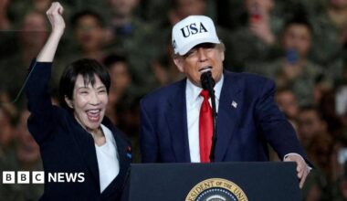 US President Donald Trump speaks as Japanese Prime Minister Sanae Takaichi visit the US' navy base in Yokosuka. Trump, wearing a white cap that reads USA, a navy suit and a red tie, has his hand on the podium while speaking. Takaichi, in a black suit, pumps her arm into the air.