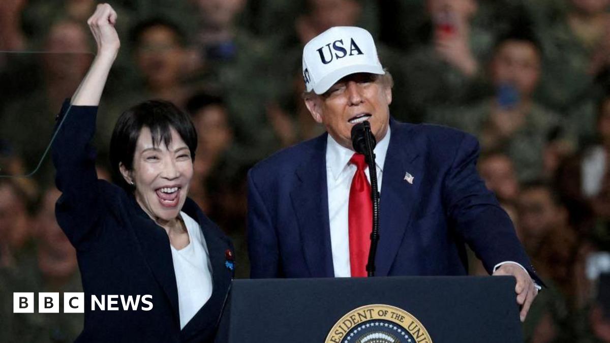 US President Donald Trump speaks as Japanese Prime Minister Sanae Takaichi visit the US' navy base in Yokosuka. Trump, wearing a white cap that reads USA, a navy suit and a red tie, has his hand on the podium while speaking. Takaichi, in a black suit, pumps her arm into the air.