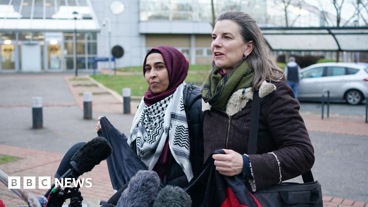 Two women standing in front of microphones outside court. The woman on the left is wearing a maroon headscard and Palestinian keffiyeh scarf around her neck. The woman on the right has shoulder length brown hair and is in a dark coat. They are about to hold up a dark banner.