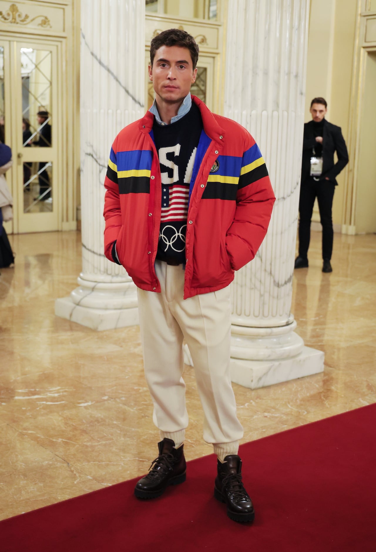 A man poses for a photo wearing a knitted navy sweater with USA on it, a red puffer jacket and cream trousers.