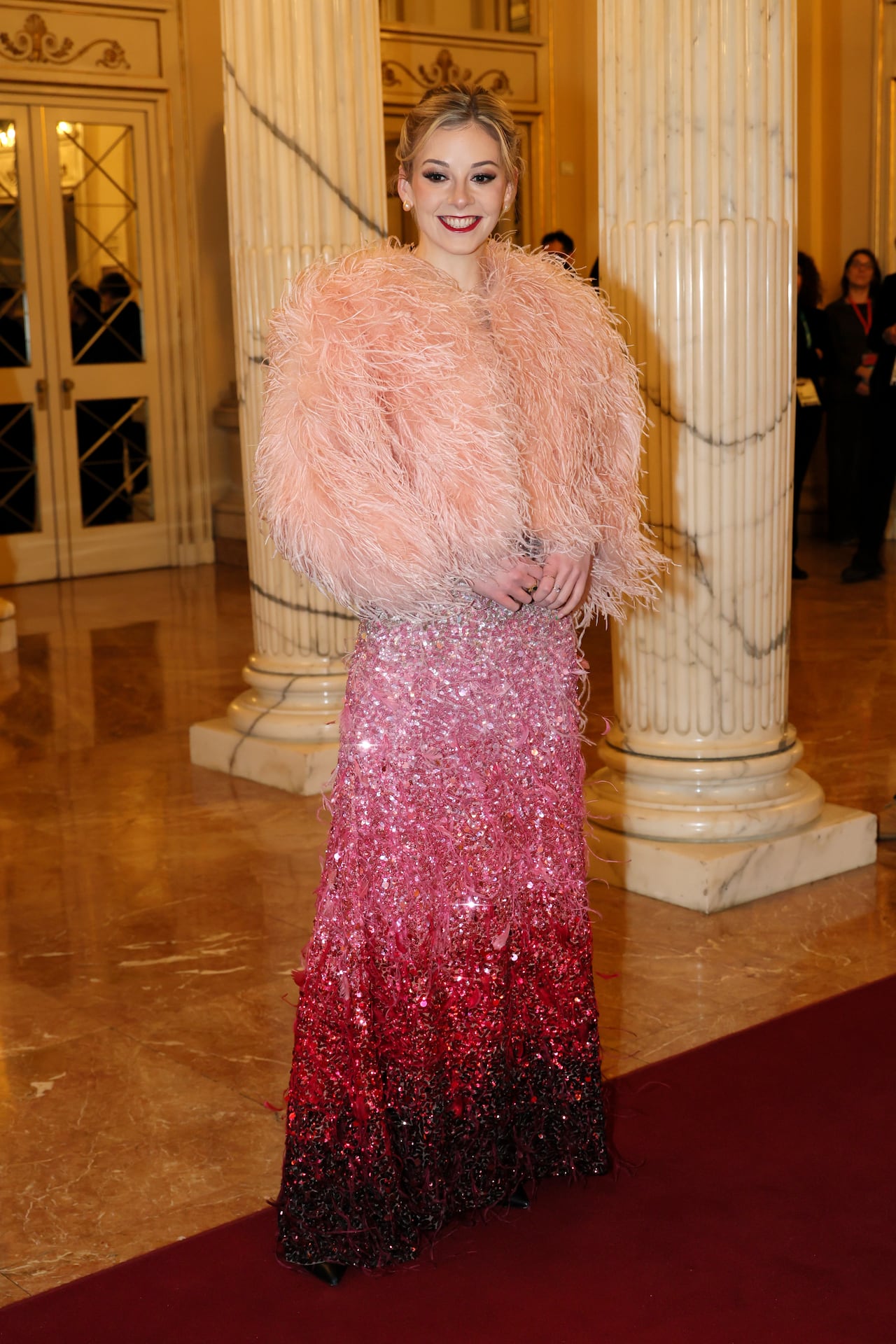A woman poses for a photo wearing a pink sequin gown with feather jacket.