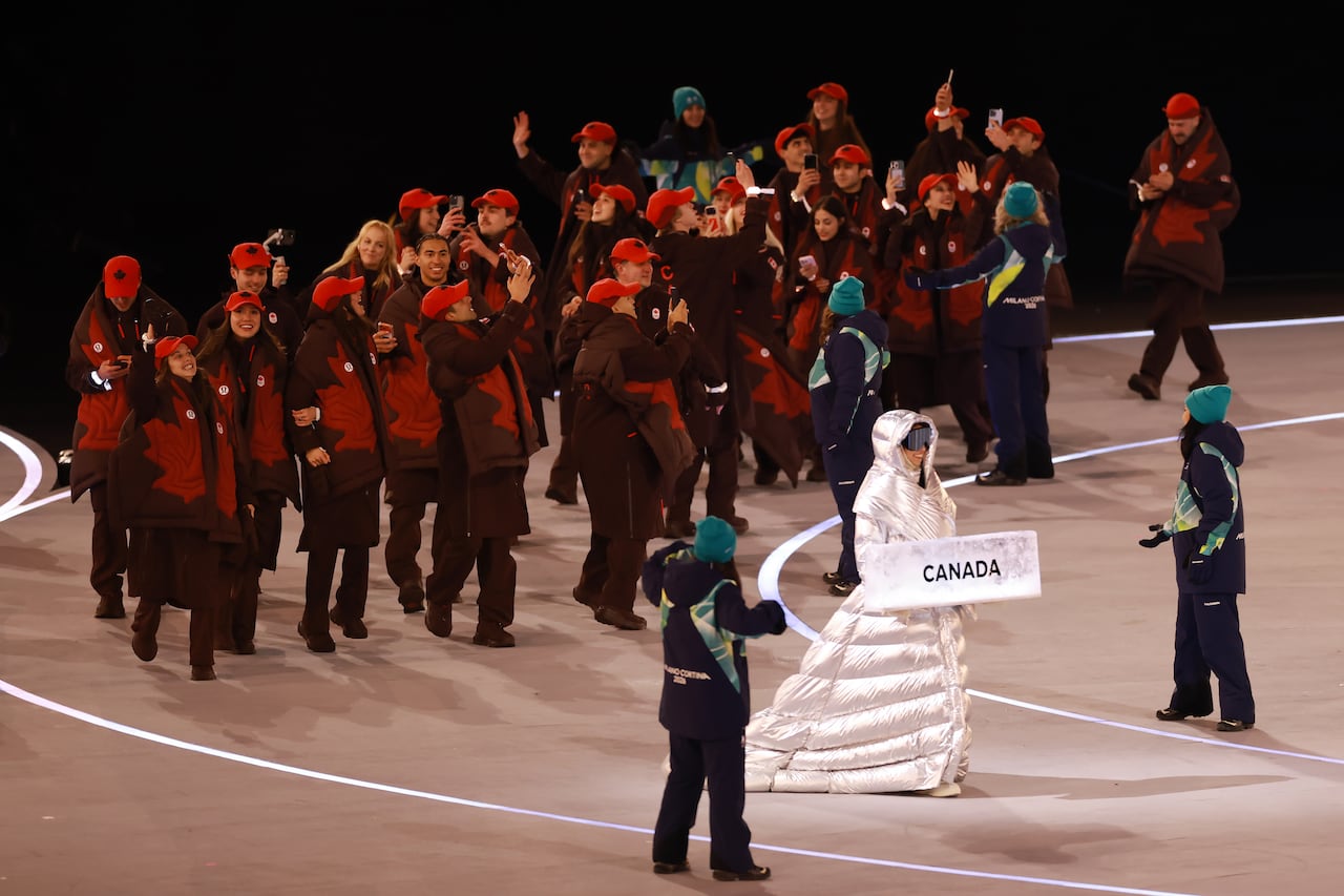 A woman wearing a silver puffer dress carries a card that says Canada with the team walking behind her. 