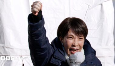Sanae Takaichi speaks into a microphone with her fist raised. She's wearing a dark blue puffer coat