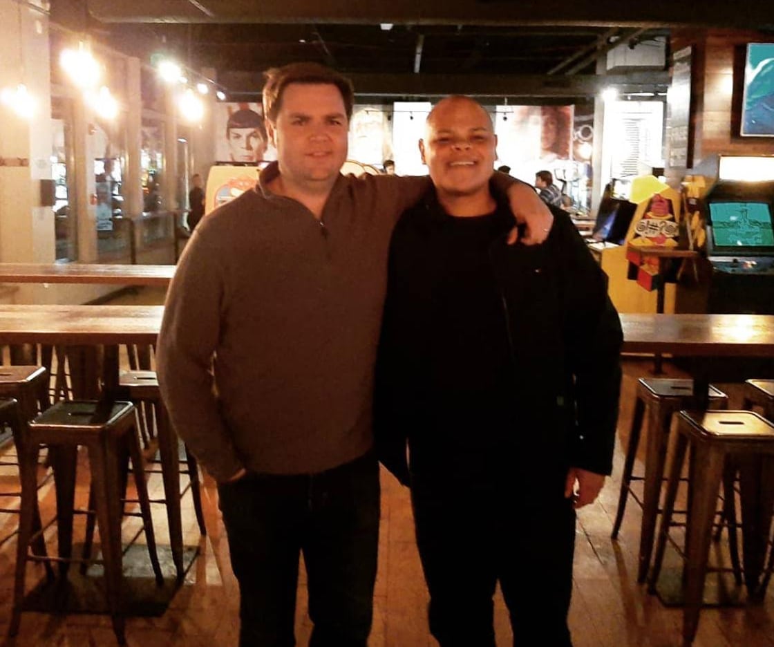 Two men stand together with one putting his arm around the other.