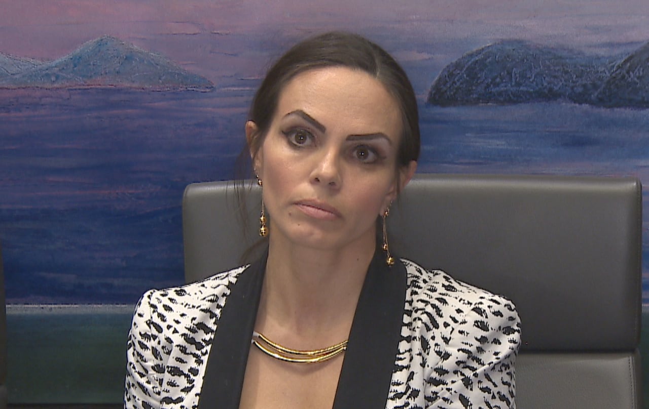 Brown-haired woman sits with frown on face in black and white blazer.