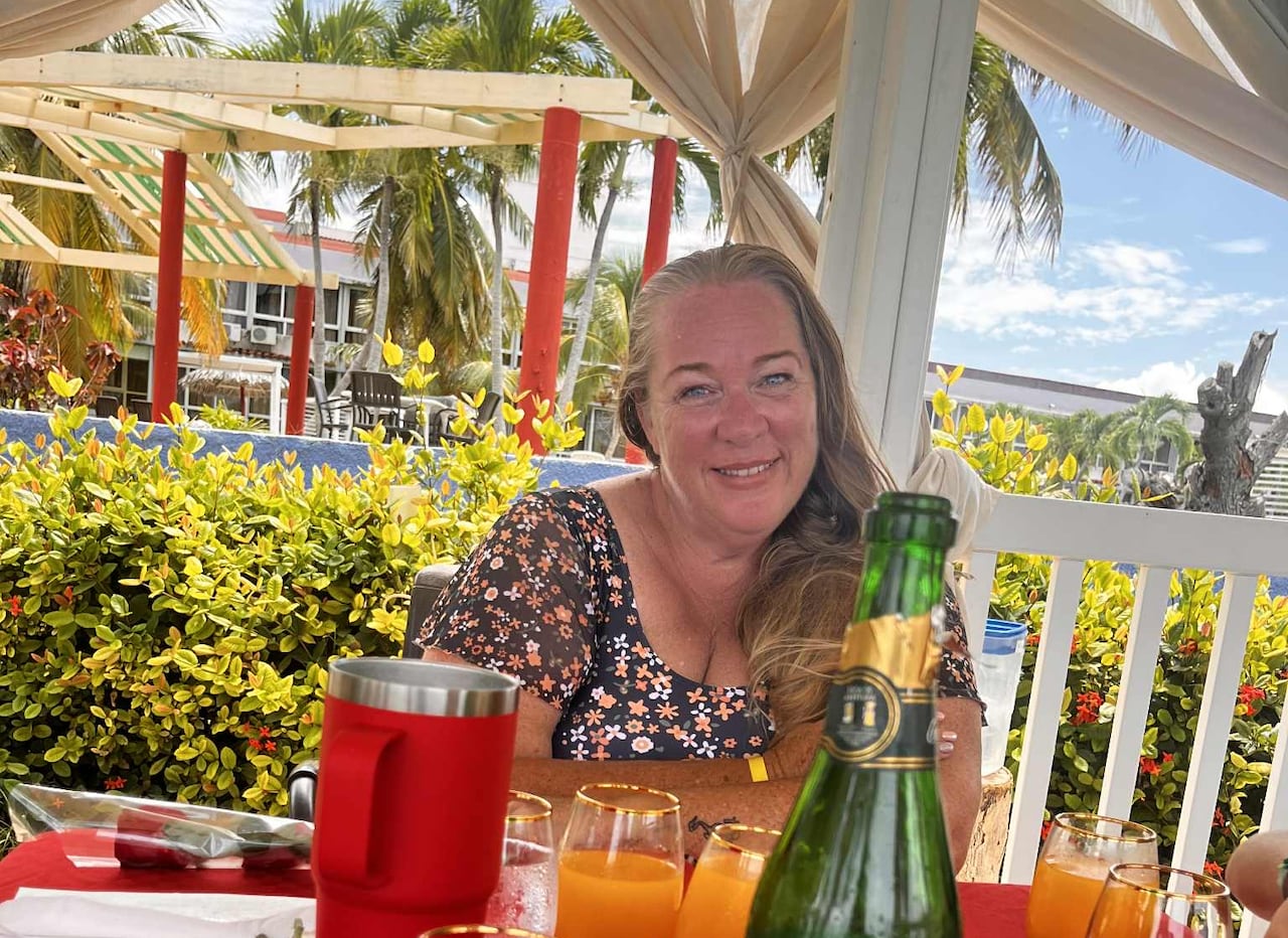 A woman smiles at a table full of glasses, in front of a tropical background