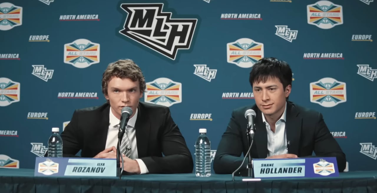 Two men sit down for a press conference. Both are wearing black blazers with white dress shirts underneath. Behind them is a series of logos 