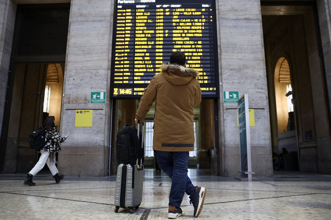 A man in a jacket holding a suitcase looks at a board announcing routes at a train station.