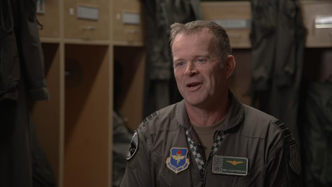 A man with short brown hair wearing an air force jumpsuit with patches and isignias speaks in a locker room setting.