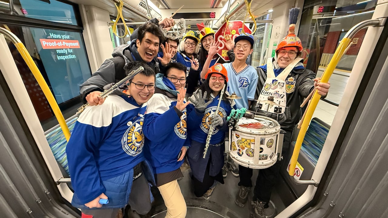 Half a dozen people wearing blue and holding musical instruments including a drum pictured aboard an interior LRT train