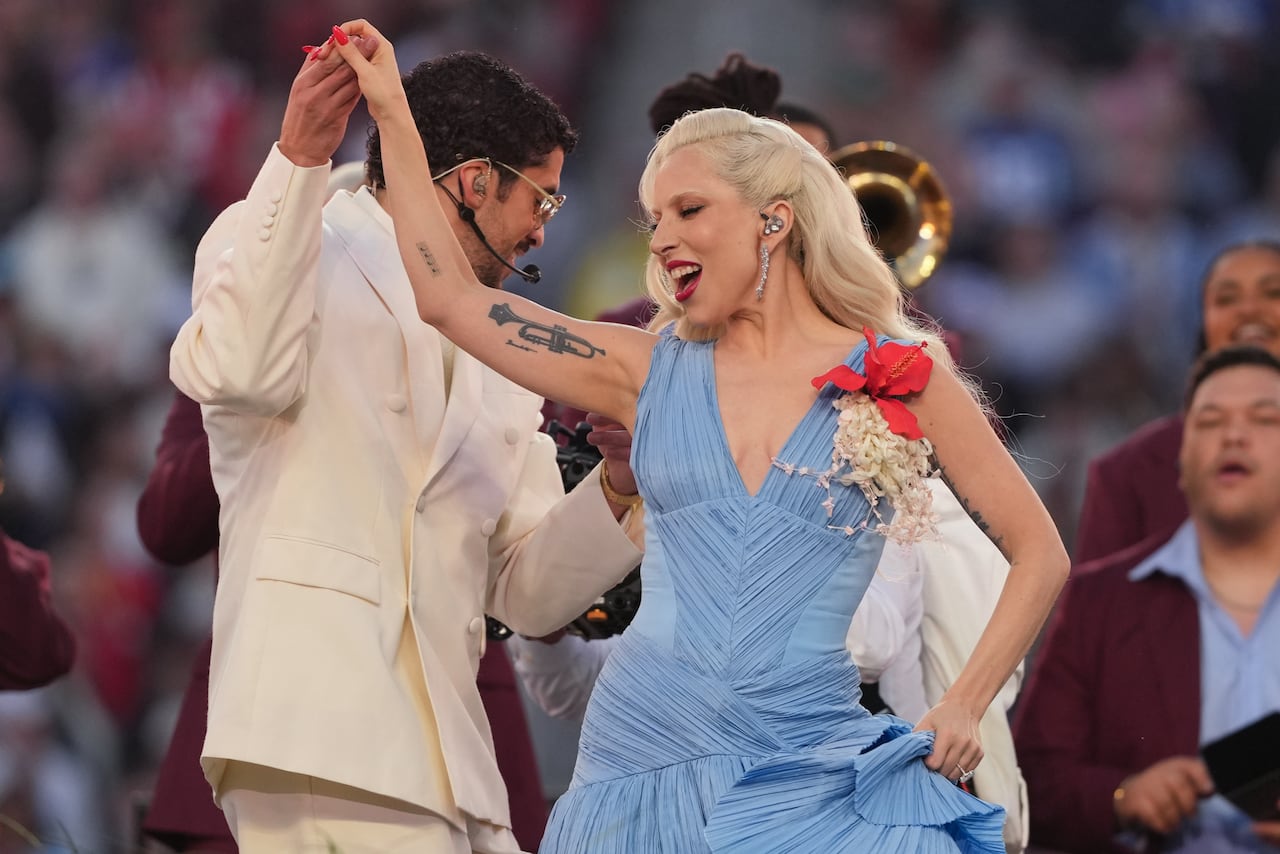 a blonde woman with a big smile who is wearing a blue salsa dress holds the hand of a man in a white suiit, as the two dance