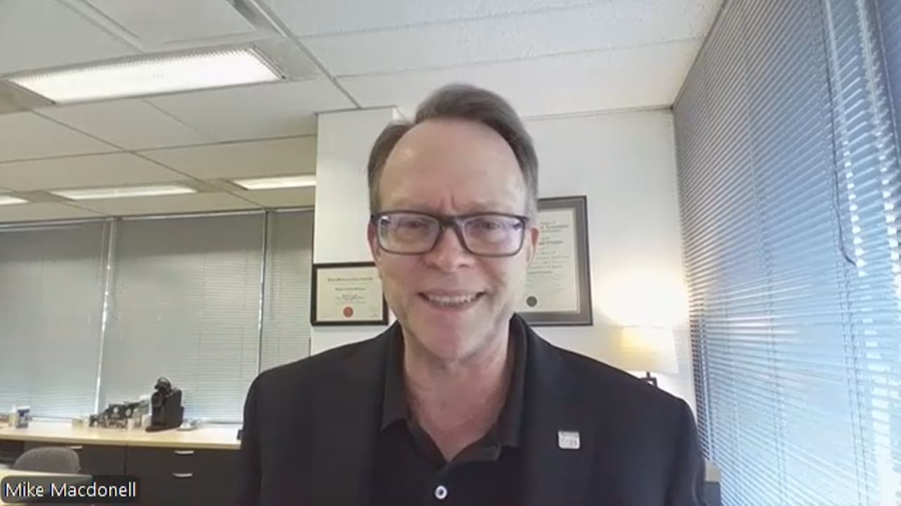 A man in suit wearing a black rimmed glass smiles during a zoom call. 
