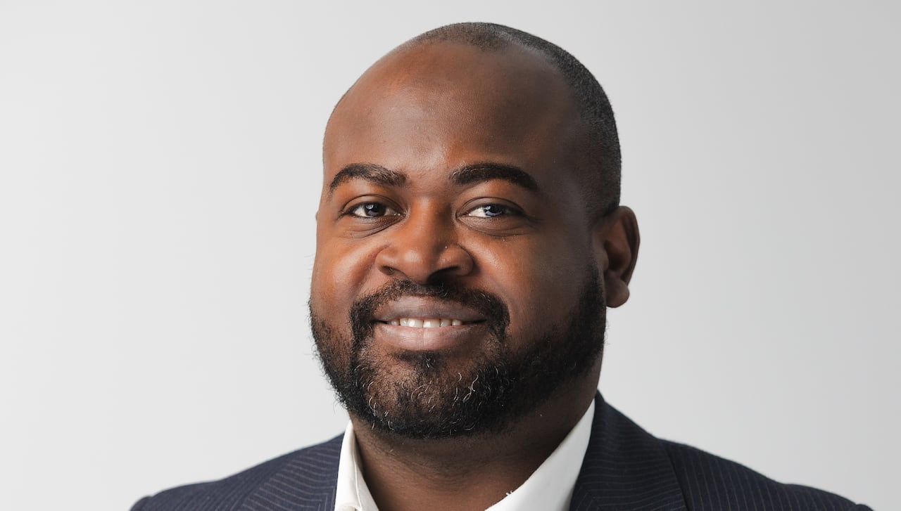 A Black man with a buzz cut and trim beard is smiling while wearing a white dress shirt and pinstripe suit jacket.