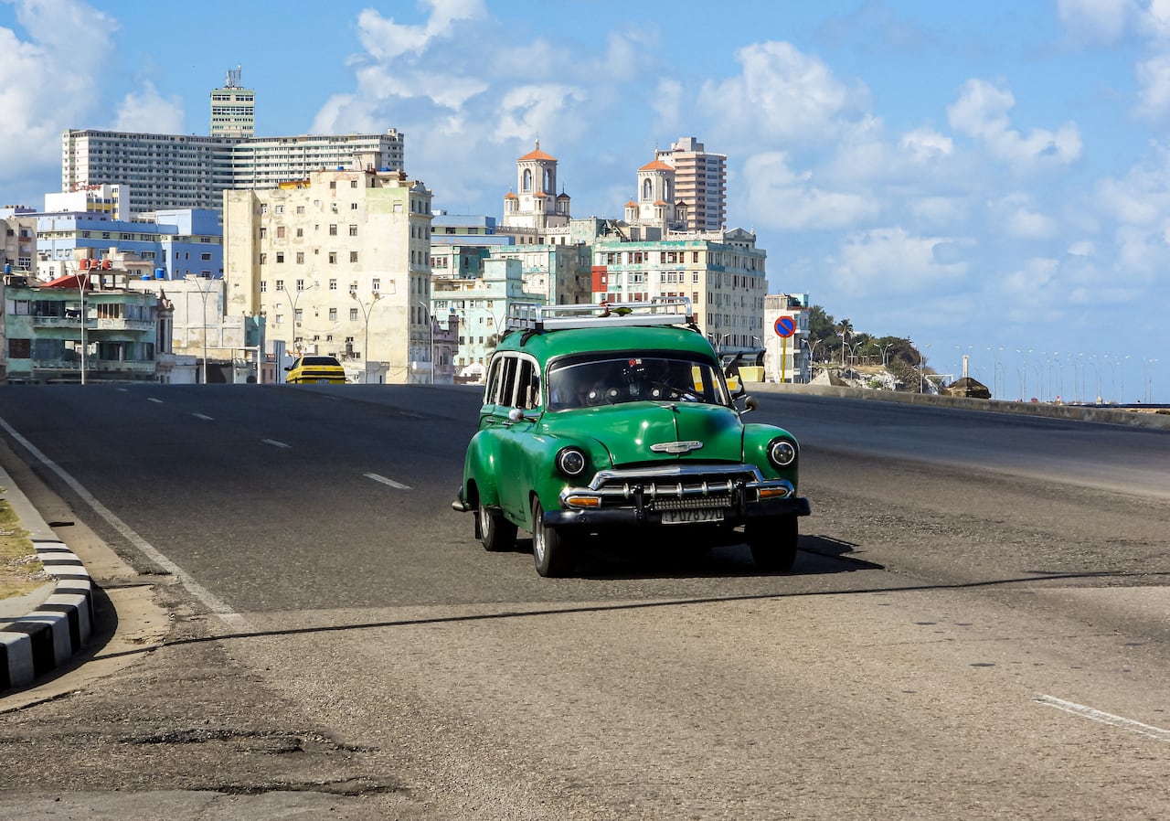 A green old car drives in front of a tropical cityscape on an empty road