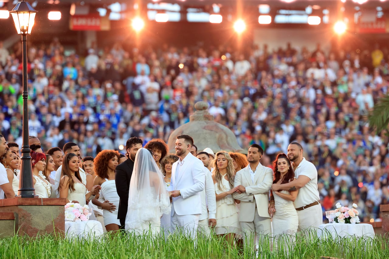 a man and woman dressed in white wedding attire look at eachother and smile, surrounded by a crowd at a wedding ceremony. in the background, pieces of the set of a musical performance and a large crowd are visible