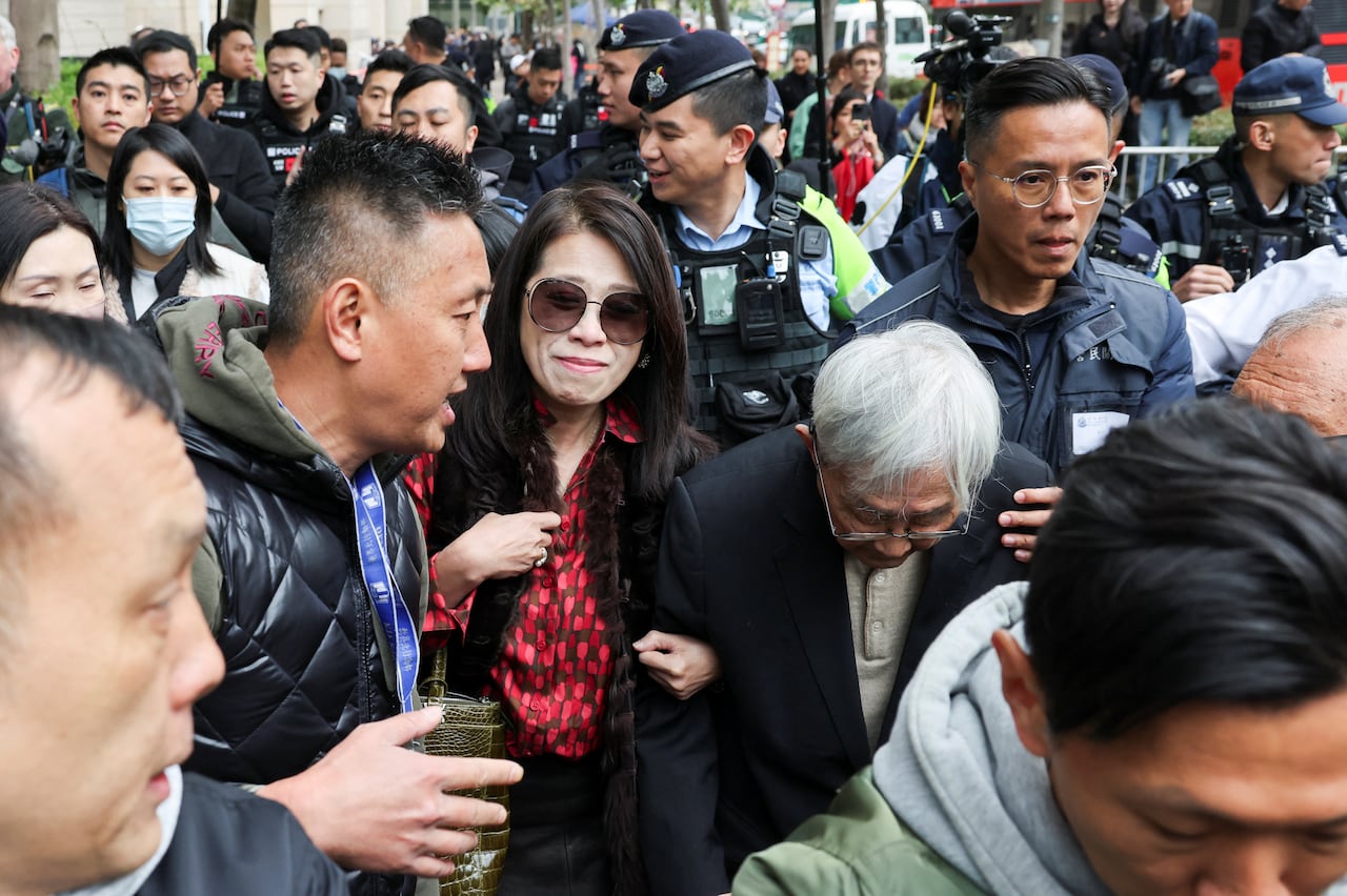 A crowd of people is shown outdoors, with the focus on a dark haired woman wearing a red top and sunglasses.