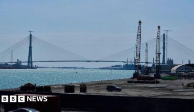 Street in Concordia, Mexico. A car and several people are in the foreground, with houses and trees on a gill in the background
