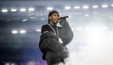 A performer wearing a dark beanie, oversized furry jacket, and a large chain necklace holds a microphone, surrounded by stage fog and bright lights.