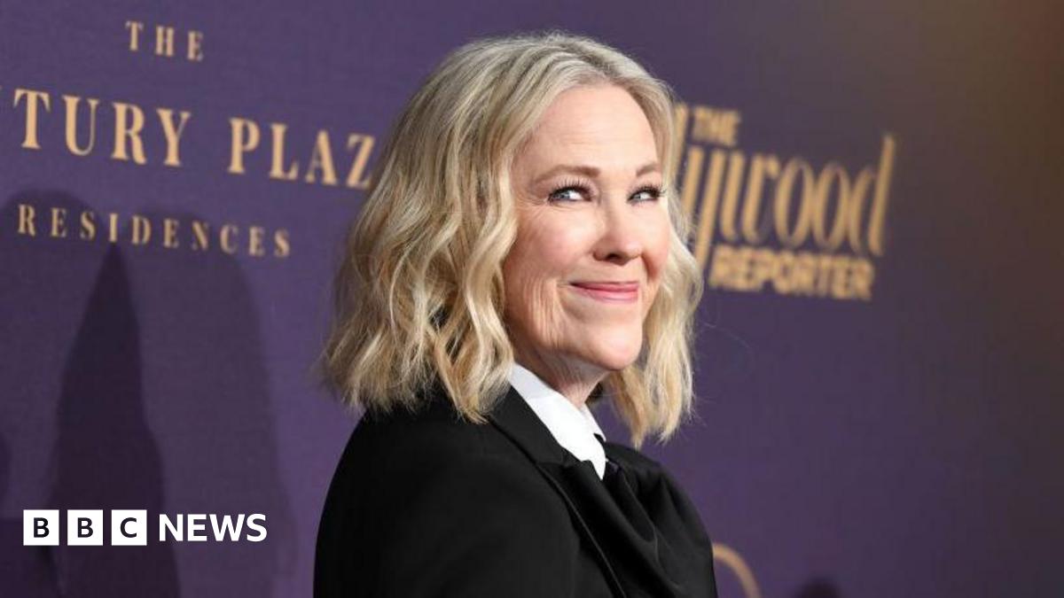 A woman with shoulder-length blonde hair is pictured in front of a purple backdrop. She wears a black suit jacket and a white shirt.