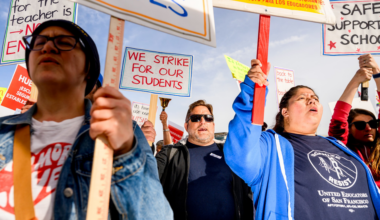 A performer wearing a dark beanie, oversized furry jacket, and a large chain necklace holds a microphone, surrounded by stage fog and bright lights.