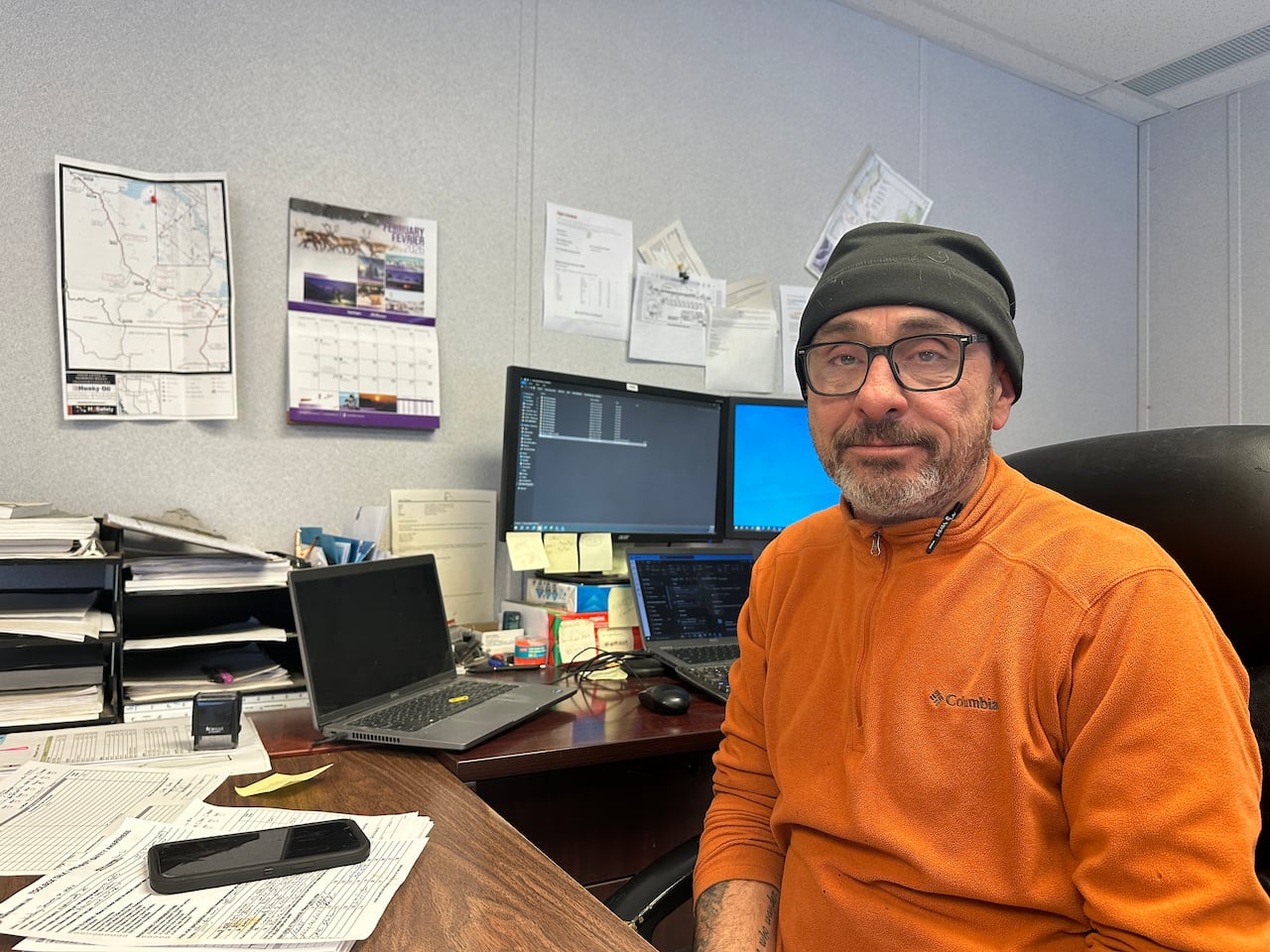 A man with a beard, orange shirt, dark toque, in an office.